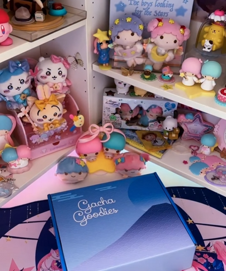 A blue Gacha Goodies gift box featuring a stylized mountain and wave design. The background is a white shelving unit filled with a diverse array of pastel-colored anime figurines and plush characters, including Pompompurin and Little Twin Stars, creating a nostalgic "dreamy" aesthetic.