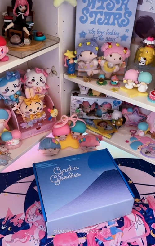 A blue Gacha Goodies gift box featuring a stylized mountain and wave design. The background is a white shelving unit filled with a diverse array of pastel-colored anime figurines and plush characters, including Pompompurin and Little Twin Stars, creating a nostalgic "dreamy" aesthetic.