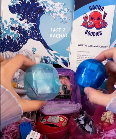 A person's hands holding two translucent blue Gachapon capsules over a "Gacha Goodies" subscription box filled with Japanese toys, including a miniature purple backpack and a toy train, set against a Great Wave-inspired background.