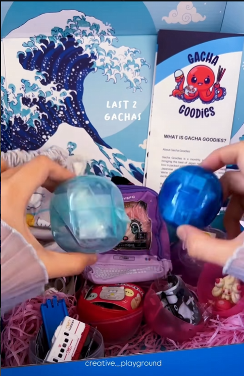 A person's hands holding two translucent blue Gachapon capsules over a "Gacha Goodies" subscription box filled with Japanese toys, including a miniature purple backpack and a toy train, set against a Great Wave-inspired background.