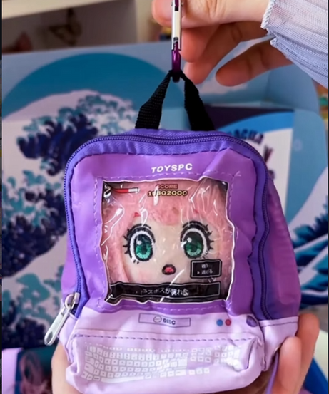 A hand holds a miniature purple "ita-bag" style backpack keychain featuring a clear window designed to look like a retro computer monitor. Inside the window is a plush pink-haired anime character, while the bottom pocket is printed with a white keyboard graphic. The keychain is attached with a purple carabiner and set against a blue Gacha Goodies box featuring a Great Wave design.