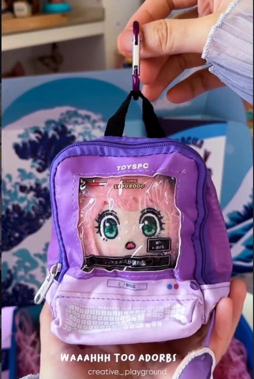A hand holds a miniature purple "ita-bag" style backpack keychain featuring a clear window designed to look like a retro computer monitor. Inside the window is a plush pink-haired anime character, while the bottom pocket is printed with a white keyboard graphic. The keychain is attached with a purple carabiner and set against a blue Gacha Goodies box featuring a Great Wave design.
