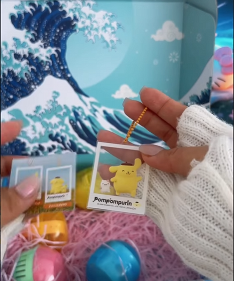 A close-up shot of hands holding an acrylic Pompompurin keychain designed to look like a Polaroid instant photo. The character Pompompurin is shown with a small companion against a yellowish background. In the background, a light blue Gacha Goodies box with a "Great Wave" illustration is visible, containing pink crinkle paper and various colorful gachapon capsules.