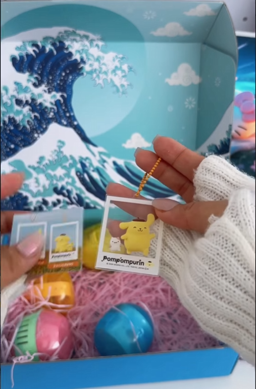 A close-up shot of hands holding an acrylic Pompompurin keychain designed to look like a Polaroid instant photo. The character Pompompurin is shown with a small companion against a yellowish background. In the background, a light blue Gacha Goodies box with a "Great Wave" illustration is visible, containing pink crinkle paper and various colorful gachapon capsules.