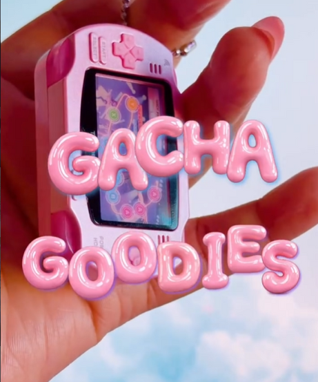 A close-up shot of a hand holding a miniature pink handheld gaming console keychain. The word "GACHA GOODIES" is overlaid in 3D pink bubble letters. The background features a soft blue sky with white clouds.