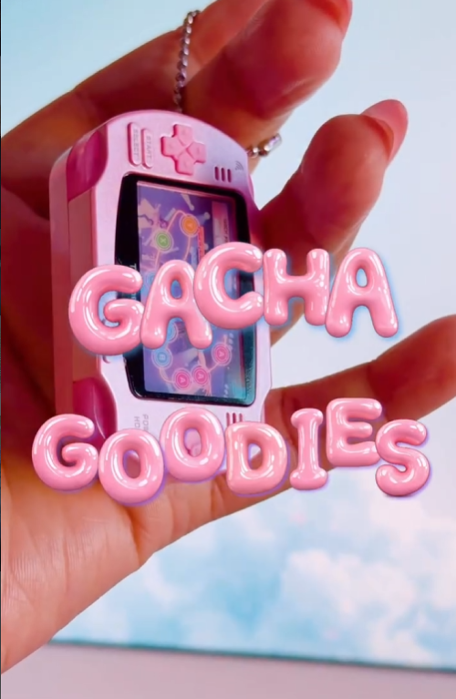 A close-up shot of a hand holding a miniature pink handheld gaming console keychain. The word "GACHA GOODIES" is overlaid in 3D pink bubble letters. The background features a soft blue sky with white clouds.