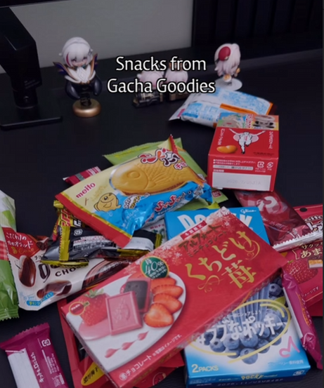 A vibrant collection of Japanese treats including chocolates, biscuits, and unique regional snacks. The assortment features bright red and blue packaging, with strawberry and blueberry themes prominently displayed alongside a miniature Taiyaki snack. Two small white anime figures stand behind the pile of sweets.