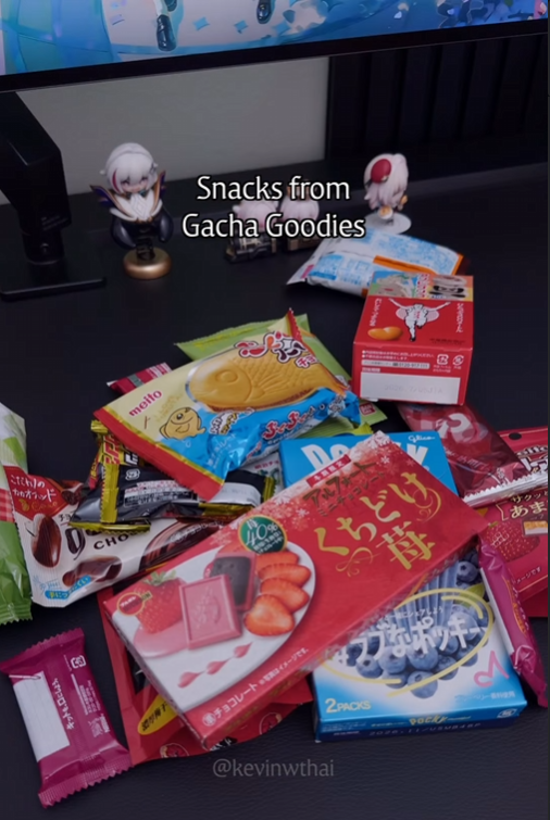 A vibrant collection of Japanese treats including chocolates, biscuits, and unique regional snacks. The assortment features bright red and blue packaging, with strawberry and blueberry themes prominently displayed alongside a miniature Taiyaki snack. Two small white anime figures stand behind the pile of sweets.